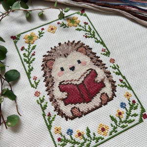 May include: Cross-stitch embroidery of a cute hedgehog reading a book, framed by a green border with flowers. The hedgehog has brown quills and is holding a red book. Embroidery floss in various colors is visible in the background.