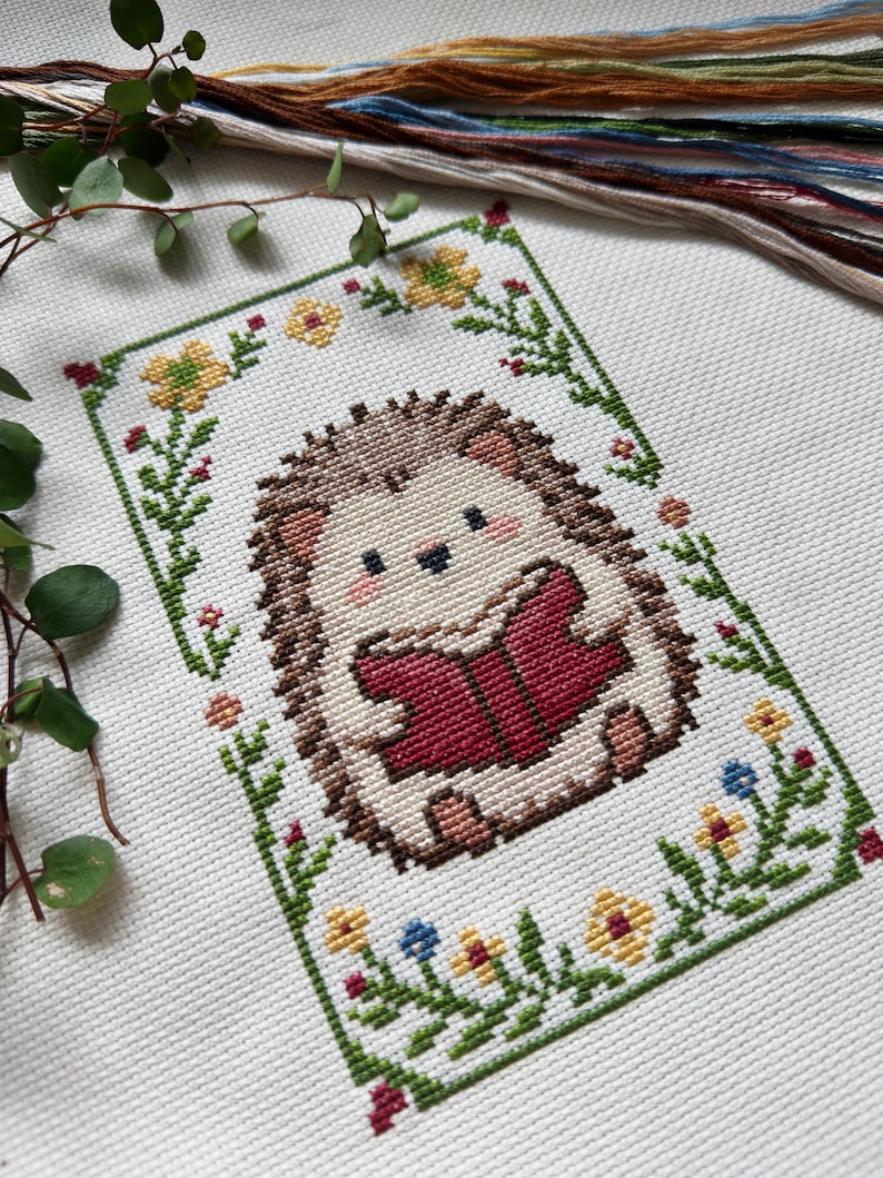 May include: Cross-stitch embroidery of a cute hedgehog reading a book, framed by a green border with flowers. The hedgehog has brown quills and is holding a red book. Embroidery floss in various colors is visible in the background.