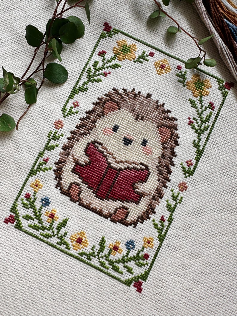 May include: Cross-stitch of a cute hedgehog reading a book, surrounded by a floral border. The hedgehog has brown quills, a cream-colored body, and is holding a red book. The border features green leaves, yellow, red, and blue flowers.