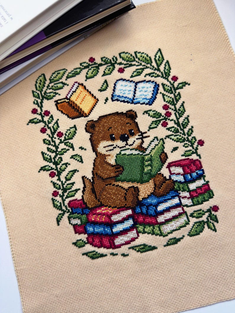 May include: Cross-stitch of a brown otter reading a green book, surrounded by a green and red floral arch. The otter sits on a stack of colorful books. Two books float above the otter. The background is a beige fabric.
