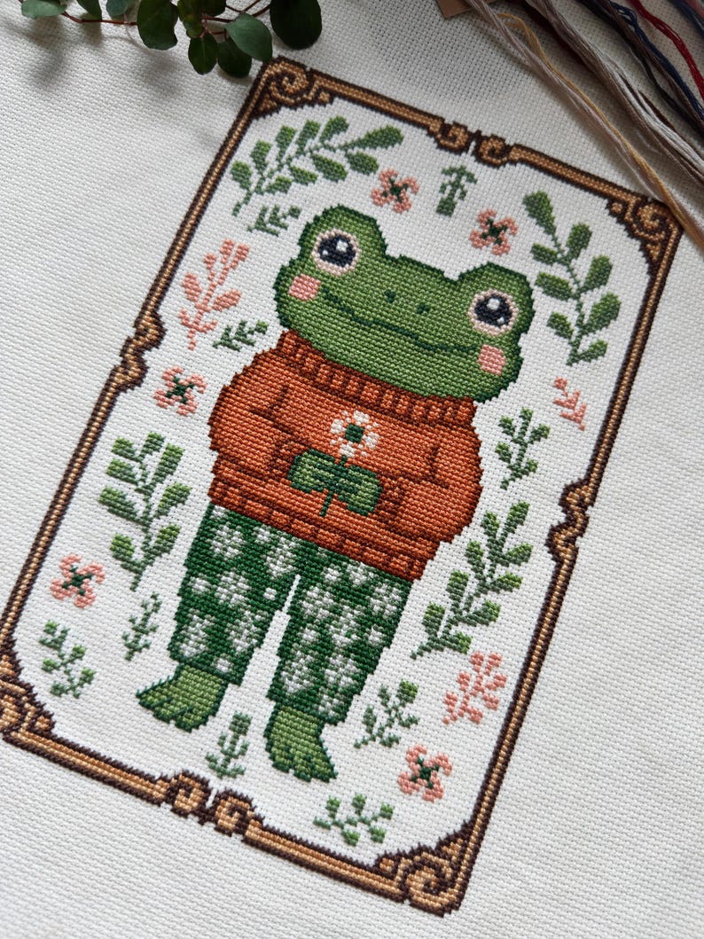 May include: Cross-stitch design of a green frog wearing an orange sweater and green patterned pants. The frog is surrounded by floral and leaf motifs, framed by a decorative border. The design is on a white fabric background.