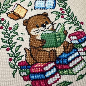 May include: Cross-stitch design featuring a brown otter reading a green book while sitting on a stack of colorful books. The design includes floating books and a green and red floral border, all on a beige background.