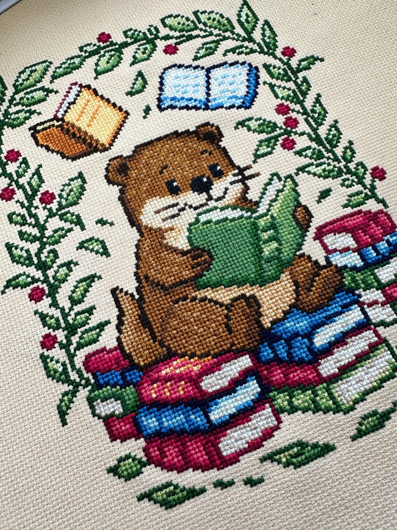 May include: Cross-stitch design featuring a brown otter reading a green book while sitting on a stack of colorful books. The design includes floating books and a green and red floral border, all on a beige background.
