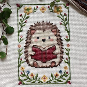May include: Cross-stitch embroidery of a hedgehog reading a book, surrounded by a floral border. The hedgehog has brown quills, a cream-colored face, and is holding a red book. The border features green leaves, yellow and blue flowers, and red accents.
