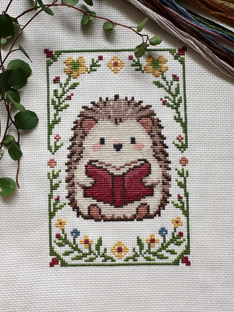 May include: Cross-stitch embroidery of a hedgehog reading a book, surrounded by a floral border. The hedgehog has brown quills, a cream-colored face, and is holding a red book. The border features green leaves, yellow and blue flowers, and red accents.