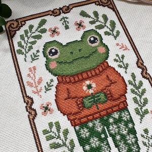 May include: Cross-stitch pattern of a frog wearing an orange sweater and green patterned pants, holding a flower. The frog has large eyes and is surrounded by floral and leaf motifs in green, pink, and brown. The design is framed with a decorative border.