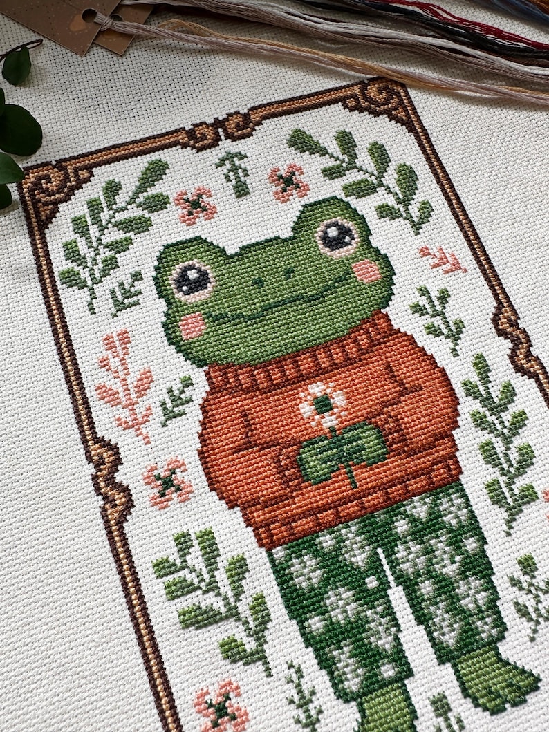 May include: Cross-stitch pattern of a frog wearing an orange sweater and green patterned pants, holding a flower. The frog has large eyes and is surrounded by floral and leaf motifs in green, pink, and brown. The design is framed with a decorative border.