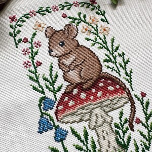 Puede incluir: Bordado de punto de cruz de un rat&oacute;n marr&oacute;n sentado sobre una seta roja y blanca, rodeado de follaje verde, flores azules y un borde floral. El fondo es blanco.