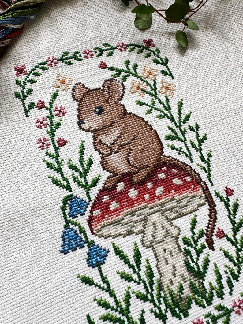 Puede incluir: Bordado de punto de cruz de un rat&oacute;n marr&oacute;n sentado sobre una seta roja y blanca, rodeado de follaje verde, flores azules y un borde floral. El fondo es blanco.