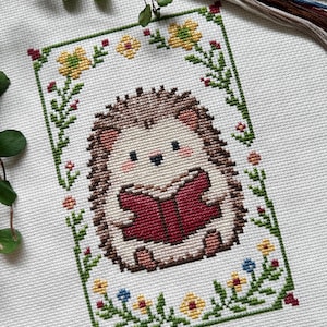 May include: Cross-stitch of a cute hedgehog reading a book, surrounded by a floral border. The hedgehog has brown quills, a cream-colored face, and is holding a red book. The border features yellow, blue, and red flowers with green leaves.