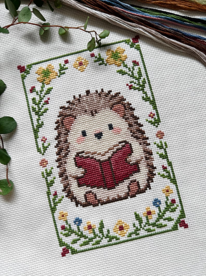 May include: Cross-stitch of a cute hedgehog reading a book, surrounded by a floral border. The hedgehog has brown quills, a cream-colored face, and is holding a red book. The border features yellow, blue, and red flowers with green leaves.