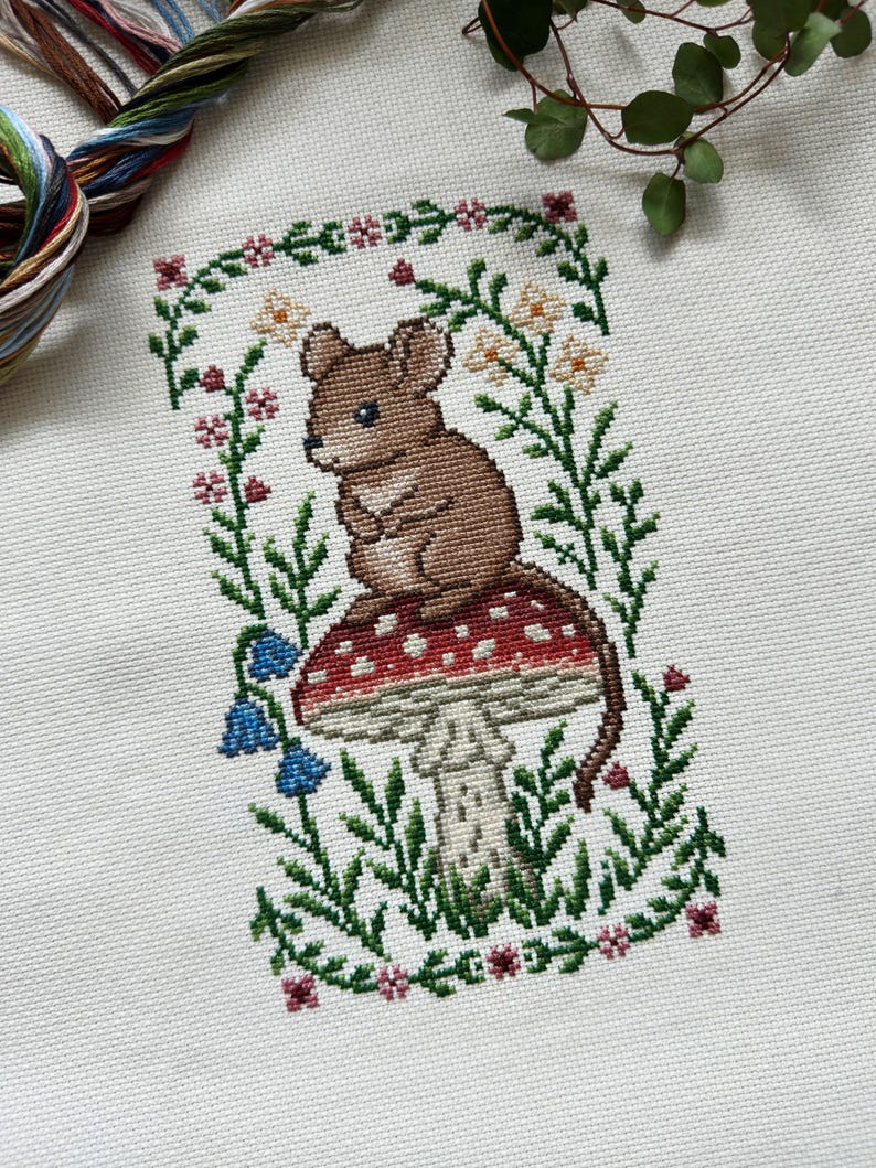 Puede incluir: Bordado a punto de cruz de un rat&oacute;n marr&oacute;n sentado sobre una seta roja y blanca, rodeado de follaje verde, flores y campanillas. El dise&ntilde;o est&aacute; enmarcado por un borde floral, una obra de arte caprichosa con tem&aacute;tica natural.