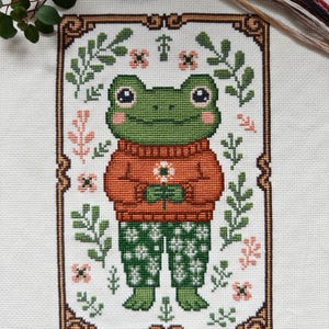 May include: Cross-stitch pattern featuring a green frog wearing an orange sweater and green patterned pants, holding a flower. The frog is framed by a brown border with floral accents and surrounded by green leaves and pink flowers. The background is white.