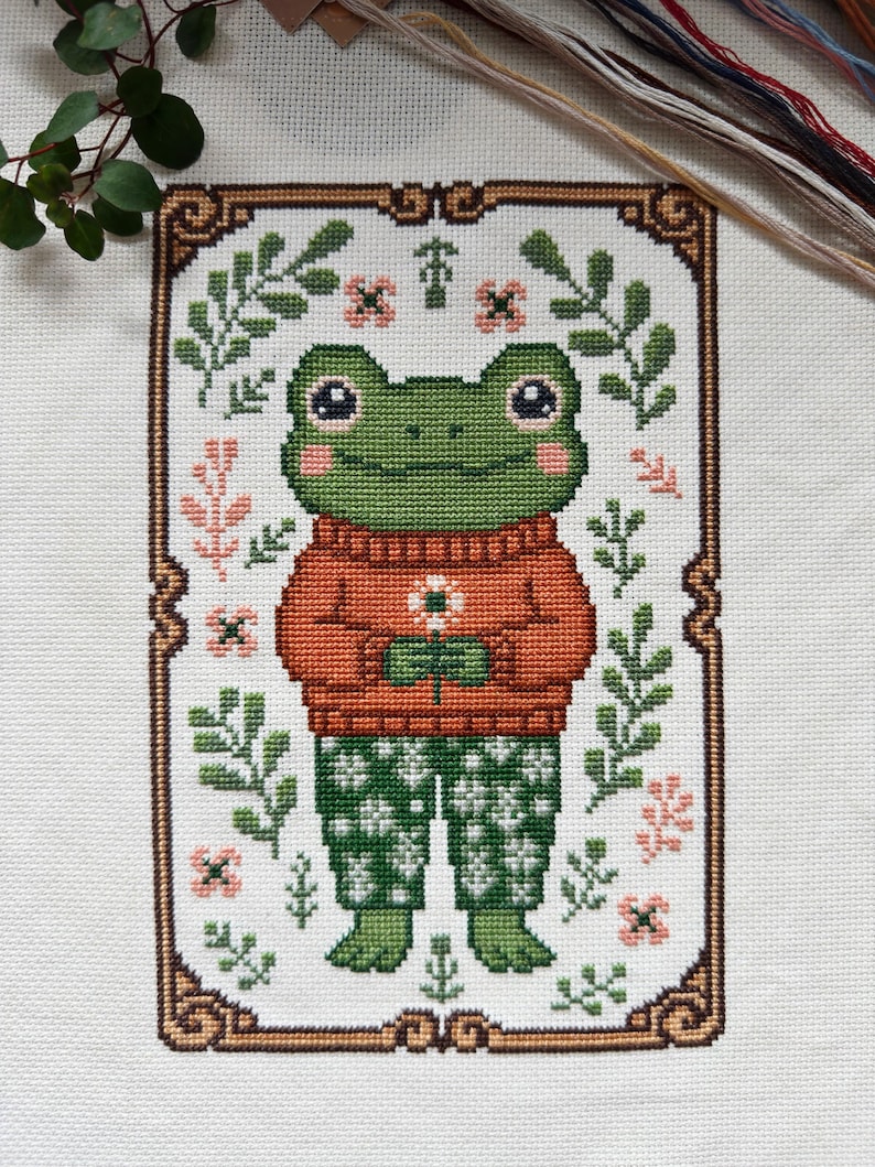 May include: Cross-stitch pattern featuring a green frog wearing an orange sweater and green patterned pants, holding a flower. The frog is framed by a brown border with floral accents and surrounded by green leaves and pink flowers. The background is white.