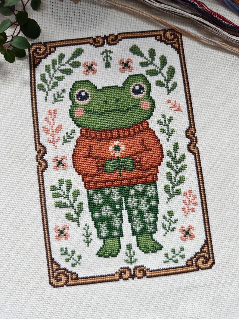 May include: Cross-stitch design featuring a green frog wearing an orange sweater and green patterned pants, holding a flower. The frog is surrounded by floral and leaf motifs, framed by a decorative border. The design is on a white background.