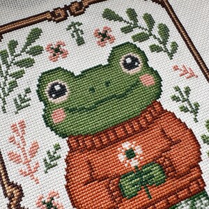 May include: Cross-stitch art featuring a green frog wearing an orange sweater with a white flower and green leaves. The frog has pink cheeks and is surrounded by floral and leaf patterns. The artwork is framed with a brown border.