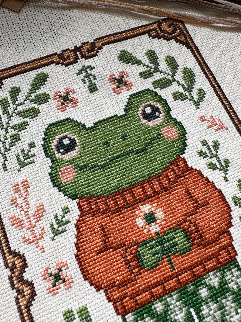 May include: Cross-stitch art featuring a green frog wearing an orange sweater with a white flower and green leaves. The frog has pink cheeks and is surrounded by floral and leaf patterns. The artwork is framed with a brown border.