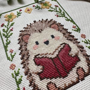 May include: A cross-stitch design featuring a cute hedgehog reading a book. The hedgehog is brown, beige, and pink, with a red book. The design is framed by green vines, yellow flowers, and red accents on a white background.