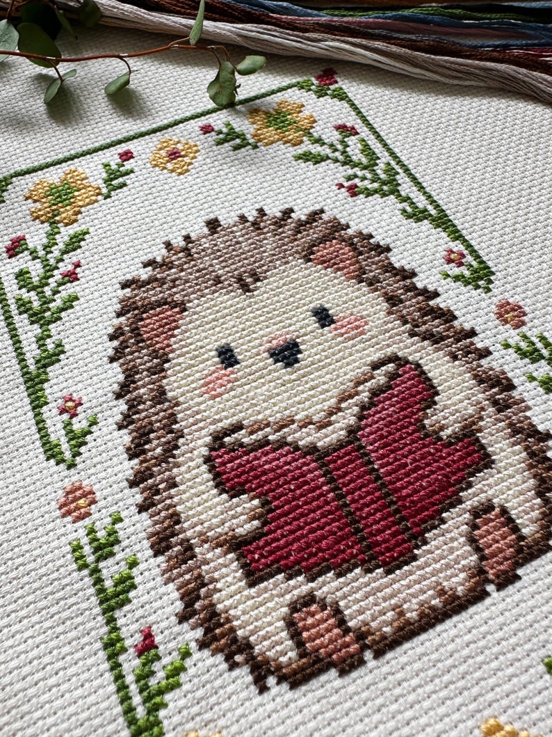 May include: A cross-stitch design featuring a cute hedgehog reading a book. The hedgehog is brown, beige, and pink, with a red book. The design is framed by green vines, yellow flowers, and red accents on a white background.