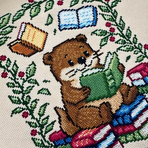 May include: Cross-stitch design featuring a brown otter reading a green book while sitting on a stack of books. The otter is surrounded by a green leafy border with red berries and two additional books. The background is a light beige color.