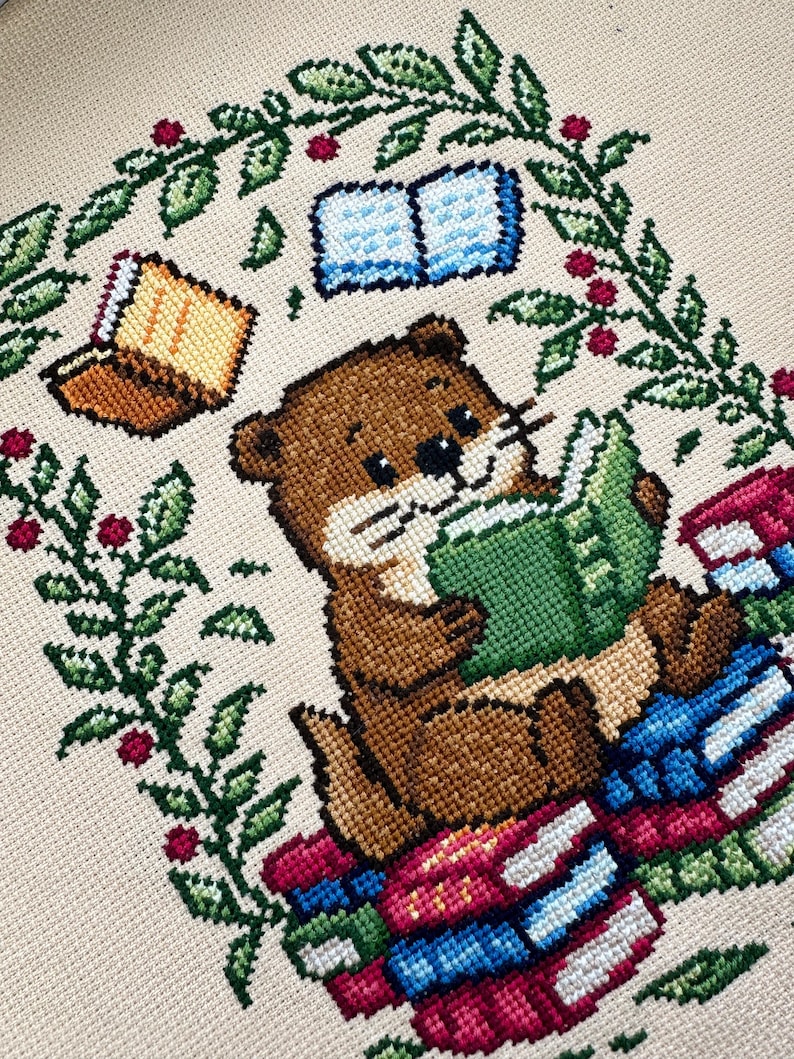 May include: Cross-stitch design featuring a brown otter reading a green book while sitting on a stack of books. The otter is surrounded by a green leafy border with red berries and two additional books. The background is a light beige color.