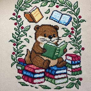 May include: Cross-stitch design featuring a brown otter reading a green book while sitting on a stack of colorful books. The otter is surrounded by a green leafy border with red berries and floating books.