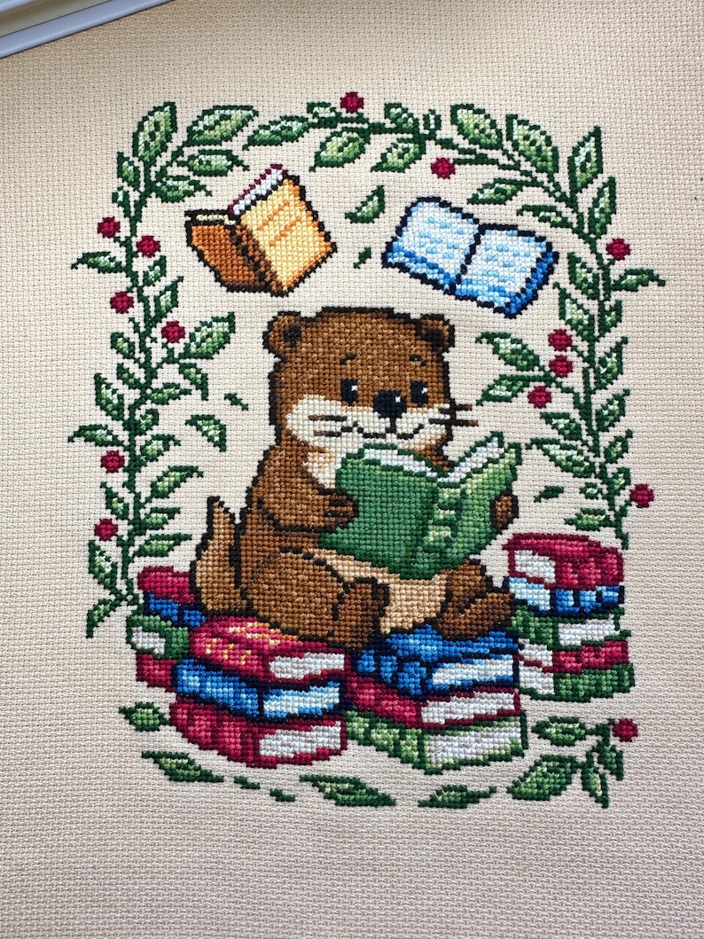 May include: Cross-stitch design featuring a brown otter reading a green book while sitting on a stack of colorful books. The otter is surrounded by a green leafy border with red berries and floating books.