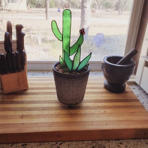 May include: A stained glass cactus with a pink flower in a gray pot with a black and white pattern. The cactus is on a wooden cutting board.