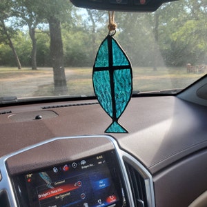 May include: A teal blue stained glass fish with a cross inside hanging from the rearview mirror of a car. The fish is a Christian symbol.