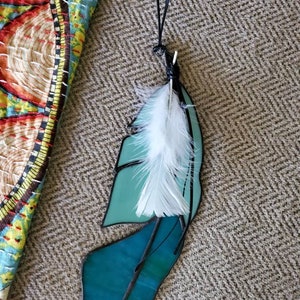 May include: Teal blue stained glass feather with a white feather accent hanging from a black cord.