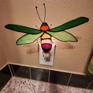 May include: A stained glass night light shaped like a colorful insect. The insect has green wings, a multi-colored body, and is plugged into a wall outlet. The light is on, emitting a soft glow.