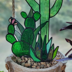 May include: A stained glass cactus plant hanging from a rope in a white ceramic pot with a textured surface. The pot has a row of small, round, orange-red accents around the bottom edge.
