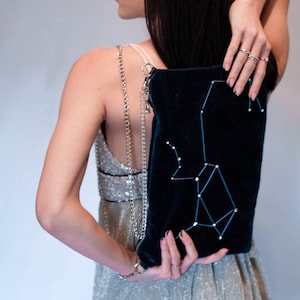 Peut inclure: Une femme portant une robe argentée à paillettes tient une pochette en velours bleu foncé avec un motif de constellation. La pochette a une bandoulière en chaîne argentée.
