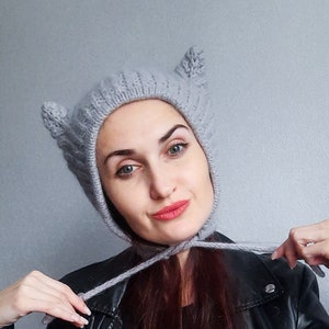 May include: A woman wearing a gray knitted hat with cat ears and a black leather jacket. The hat has a long tie that she is holding in her hands.