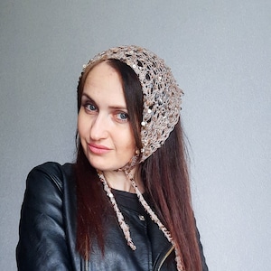 May include: A woman wearing a brown crocheted head scarf with gold sequins. The scarf has long ties that hang down her back. She is wearing a black leather jacket.