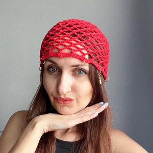 May include: A woman with long brown hair is wearing a red crocheted hat with gold rings and charms. She is looking at the camera and has her hand on her chin.