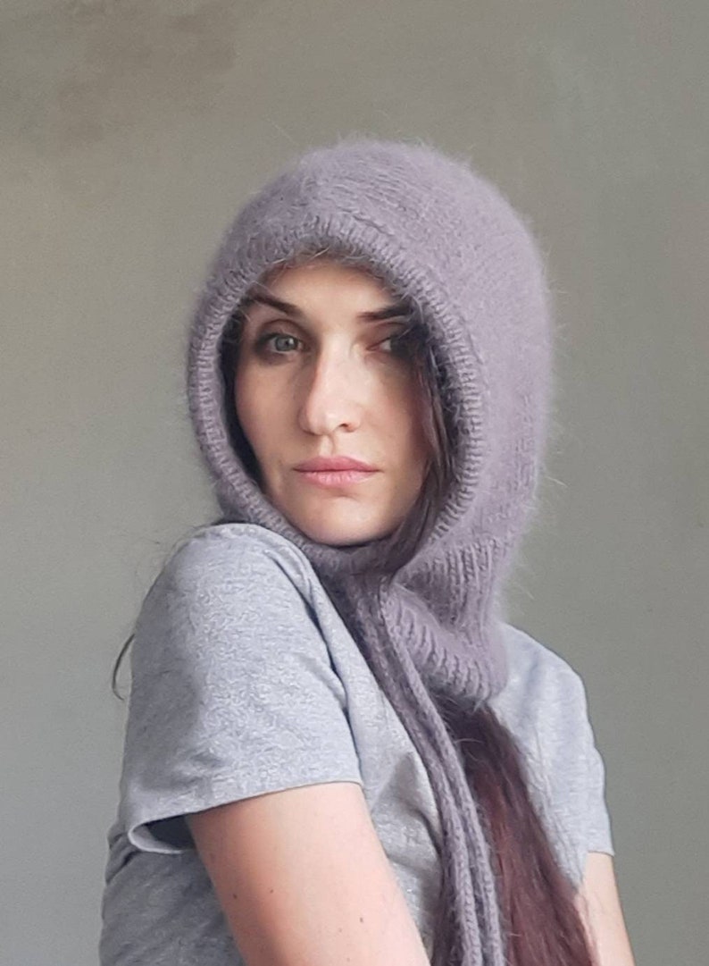 May include: A woman wearing a soft, grey hooded scarf with a long, flowing design. The scarf is made of a fuzzy, knitted material and covers her head and neck.