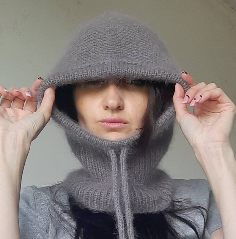 May include: A grey knitted balaclava with a long neck and a drawstring. The balaclava is made of a soft, fuzzy material.