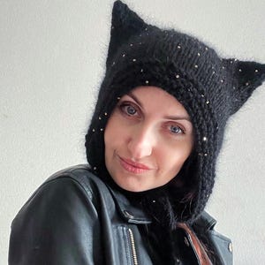 Pode incluir: Capuz preto de malha com orelhas de gato e lantejoulas prateadas. O capuz é usado sobre a cabeça e tem orelhas pontudas. A pessoa está usando uma jaqueta de couro preta.