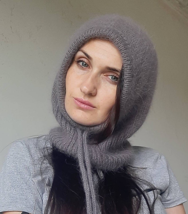 May include: A grey knitted hooded scarf with a long tassel. The scarf is made of a soft, fuzzy material.