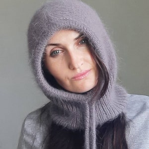 May include: A close-up of a woman wearing a soft, grey hooded cowl neck scarf. The scarf is made of a fuzzy, knitted material and has a long, flowing design.