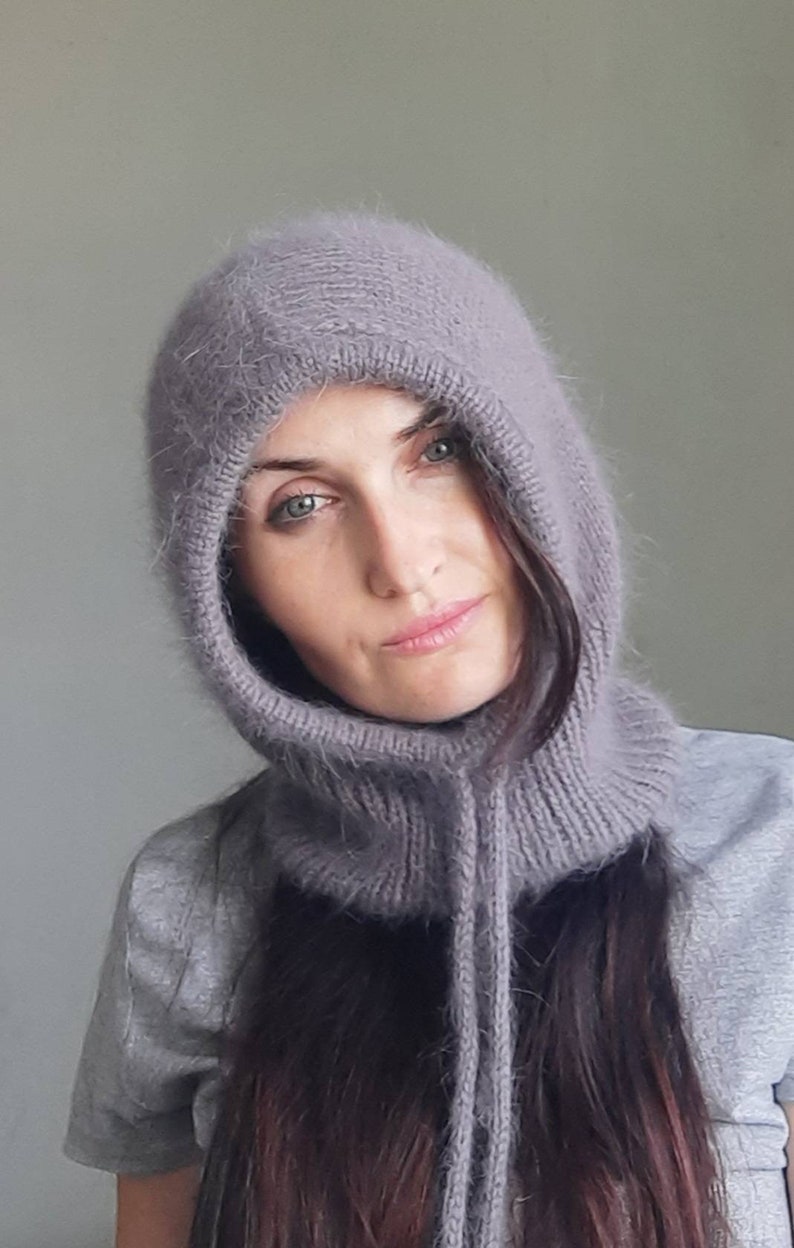 May include: A close-up of a woman wearing a soft, grey hooded cowl neck scarf. The scarf is made of a fuzzy, knitted material and has a long, flowing design.