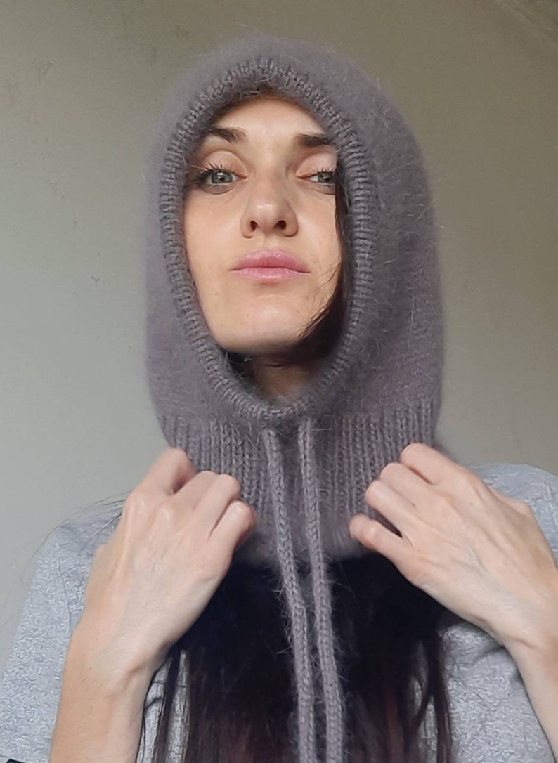 May include: A soft, grey hooded cowl with a long, fuzzy texture. The cowl has a drawstring tie at the neck.