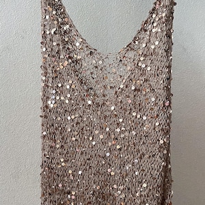 May include: A tan, open-weave top with a deep V-neck, embellished with numerous small, round, gold-colored sequins. The top is hanging on a white hanger against a neutral background. The garment appears to be lightweight.