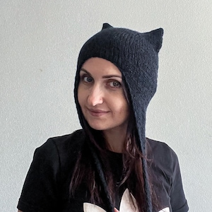 May include: A dark navy blue knit hat with cat ears. The hat covers the head and has long flaps that extend down the sides. The hat is made of a textured yarn and is worn by a person.