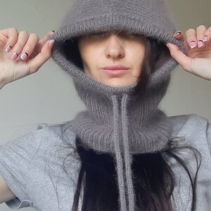 May include: A grey knitted hooded neck warmer with a long, thin drawstring. The hood is pulled up over the person's head, covering their eyes.