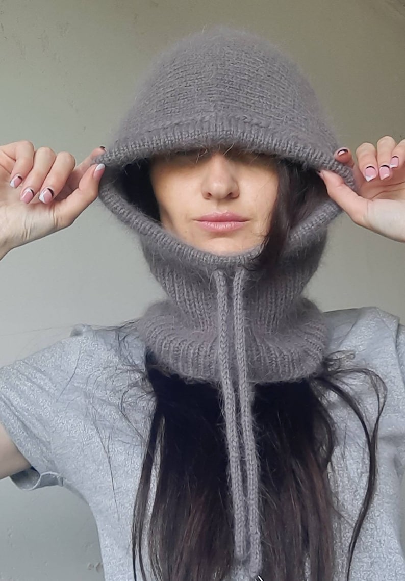 May include: A grey knitted hooded neck warmer with a long, thin drawstring. The hood is pulled up over the person's head, covering their eyes.