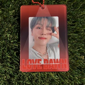 May include: A red rectangular keychain with a photo of a person winking. The keychain has the words "LOVE DAWN" in red block letters at the bottom. A red beaded chain is attached to the top.