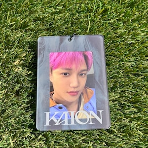 Puede incluir: Un portatarjetas fotográfico de acrílico transparente con una foto de una persona con cabello rosado. La tarjeta fotográfica tiene la palabra "KAION" impresa en blanco en la parte inferior. El fondo es verde.