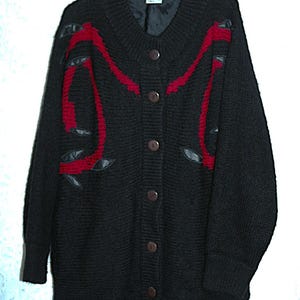 Könnte beinhalten: Schwarze Strickjacke mit Knöpfen, roten Akzenten und ledernen Blattformen. Der Pullover hat einen V-Ausschnitt und lange Ärmel. Braune Knöpfe verschließen die Vorderseite. Er hängt an einem Holzkleiderbügel.